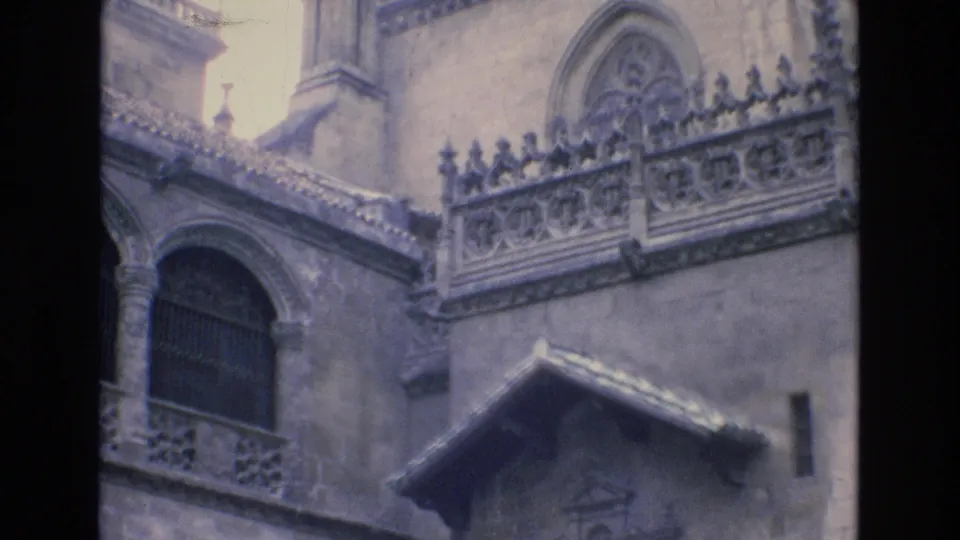 1972 Spain 8mm frame showing the upper portion of a historic stone facade, including a decorative railing, arched window, carved gable detail, and bright sky near the roofline.
