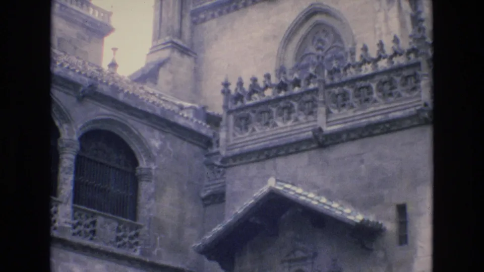 1972 Spain 8mm frame showing a historic Gothic-style stone facade with an arched barred window, ornate balustrade, and weathered roofline.