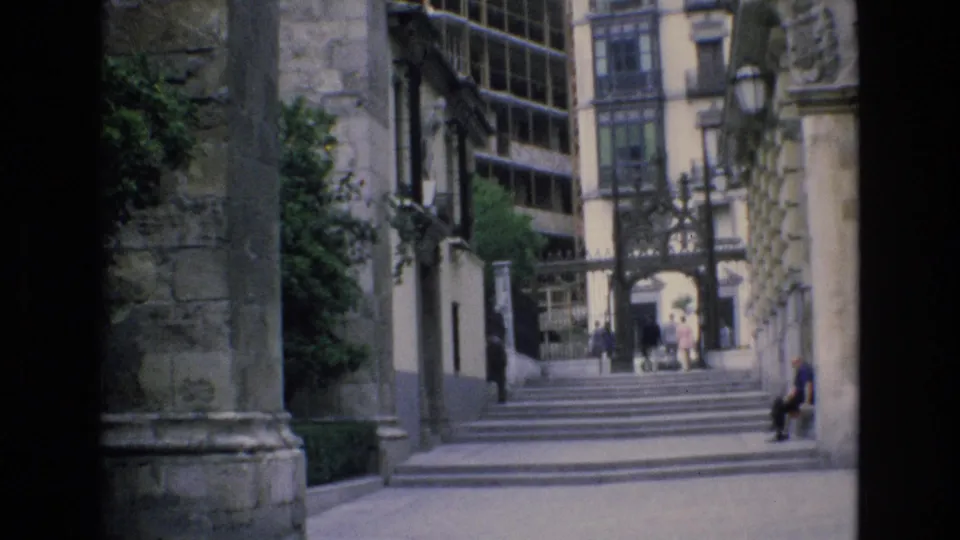 1972 Spain 8mm frame looking through a historic stone courtyard toward broad steps, an iron gate, large glazed windows, and small distant visitors.