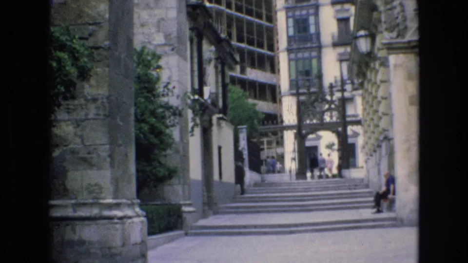 1972 Spain 8mm frame showing a historic courtyard approach with broad steps, stone walls, climbing greenery, an iron gate, and a person seated near the right edge.