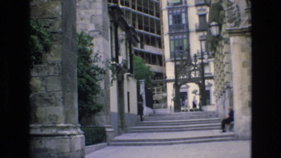 1972 Spain 8mm frame of a stone courtyard and stairway leading to an iron gate, with greenery along the wall, tall windows, distant visitors, and a seated figure.