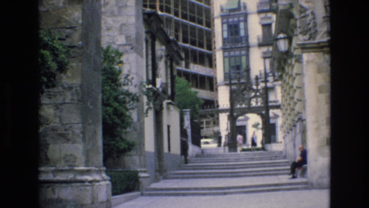 1972 Spain 8mm frame showing stone steps, old masonry, an ornate gate or archway, distant pedestrians, greenery, and a seated person at the right edge.