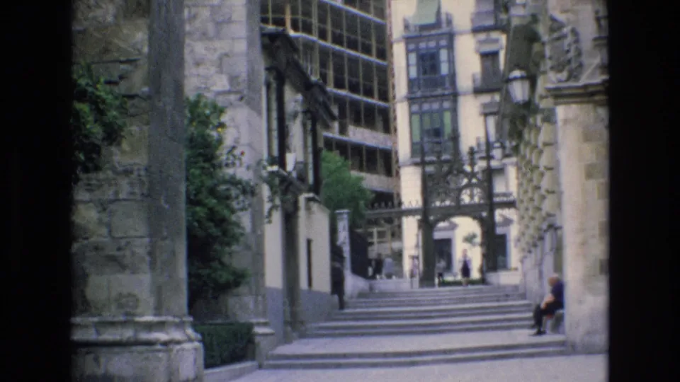 1972 Spain 8mm frame holding on stone steps framed by historic walls, with small pedestrians near the entrance and a seated figure along the right side.