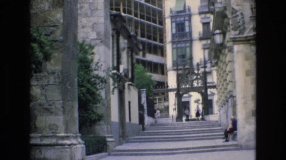 1972 Spain 8mm frame of a public stairway between old stone architecture and a modern glass facade, with distant pedestrians and a seated person at frame right.