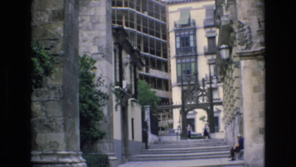1972 Spain 8mm frame showing the same urban stairway, historic stone walls, metal entrance detail, background pedestrians, and a seated person at the right edge.