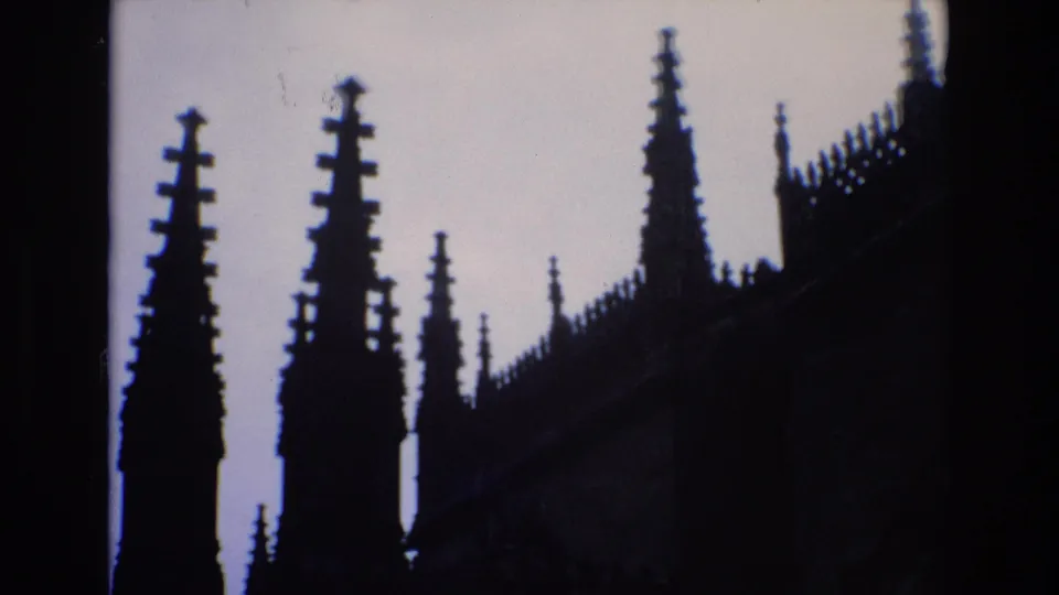 1972 Spain 8mm frame showing pointed Gothic spires and a crenellated roofline silhouetted against a pale sky, with the building mostly in shadow.