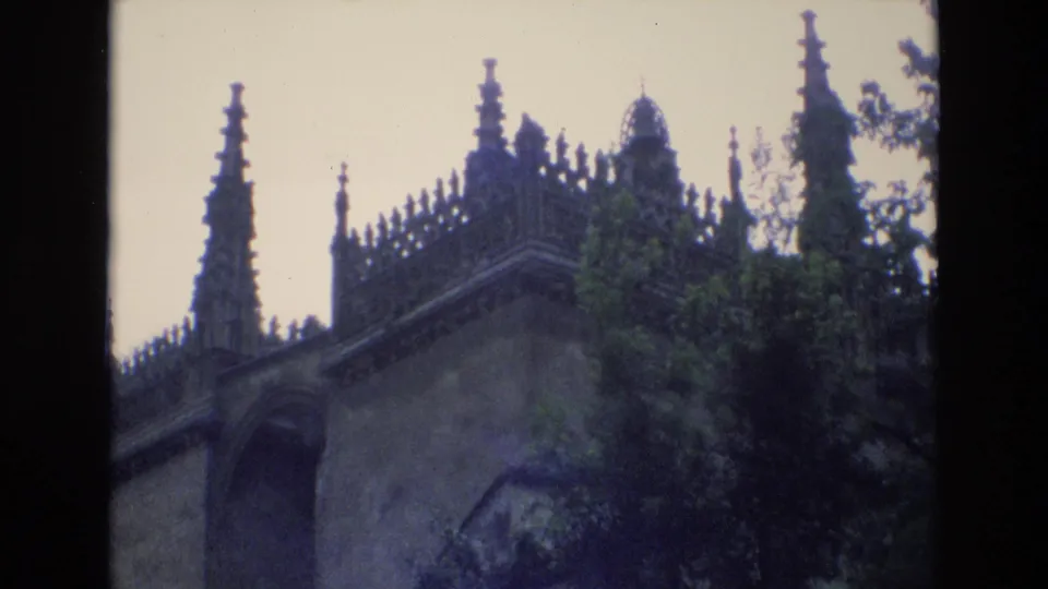 1972 Spain 8mm frame revealing more of the gray stone structure, including arched masonry, pointed spires, crenellated upper details, and leafy trees below.