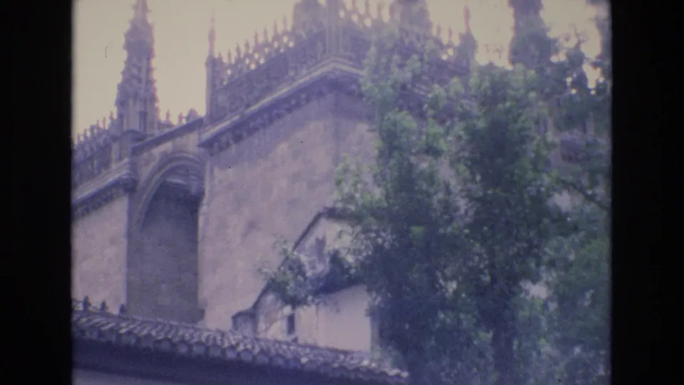1972 Spain 8mm frame showing a Gothic-style stone facade with an arched opening, decorative upper roofline, tiled lower roof edge, and green trees in the foreground.
