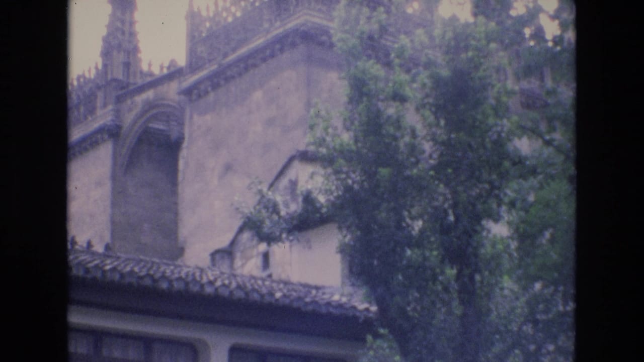 1972 Spain home-movie frame showing the upper facade of a building with a tiled roofline, arched architectural detail, and leafy trees partly obscuring the wall.