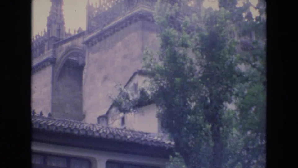 1972 Spain home-movie frame showing the upper facade of a building with a tiled roofline, arched architectural detail, and leafy trees partly obscuring the wall.