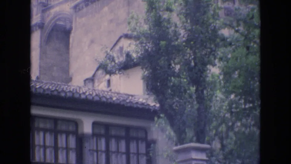 1972 Spain frame showing a building exterior with a clay tile roof, gridded windows, leafy trees, and part of a pale stone column near a fence line.
