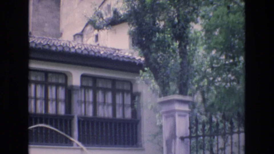 1972 Spain home-movie still showing the lower facade of a building with gridded windows, a tile roof edge, a stone column, trees, and wrought iron fencing.