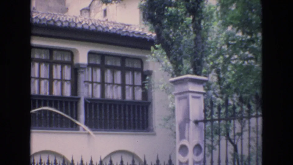 1972 Spain frame showing gridded windows and a shaded exterior wall behind a wrought iron fence, with a pale column and leafy trees in the foreground.