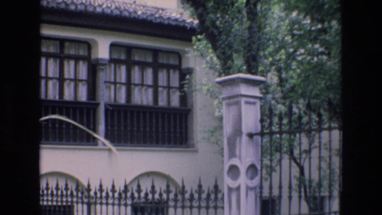 1972 Spain 8mm frame showing a cream exterior with dark window bars, arched lower openings, a tiled roof edge, trees, and an ornate iron fence beside a stone pillar.