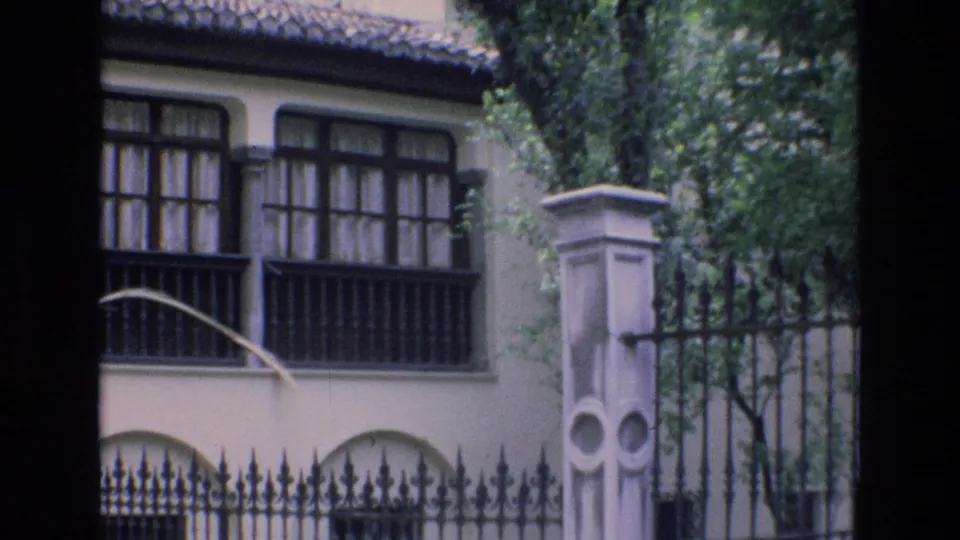 1972 Spain 8mm frame showing a cream exterior with dark window bars, arched lower openings, a tiled roof edge, trees, and an ornate iron fence beside a stone pillar.