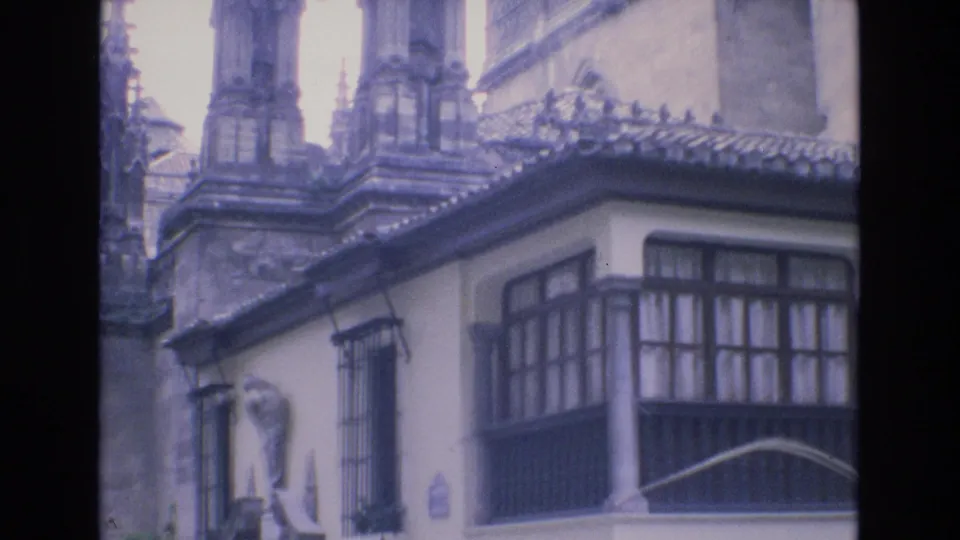1972 Spain 8mm frame showing a traditional white facade with iron window bars and wooden balcony glazing beneath Gothic cathedral stonework.