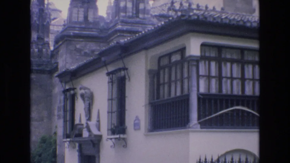 1972 Spain frame of a white stucco corner building with wooden window panels, iron bars, a small curved balcony rail, and a wall-mounted statue beneath Gothic architecture.