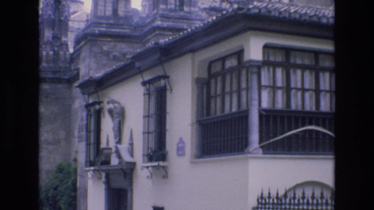 1972 Spain 8mm frame showing a pale urban building with dark balcony windows, wrought-iron grilles, a small exterior statue, and stone architecture behind it.