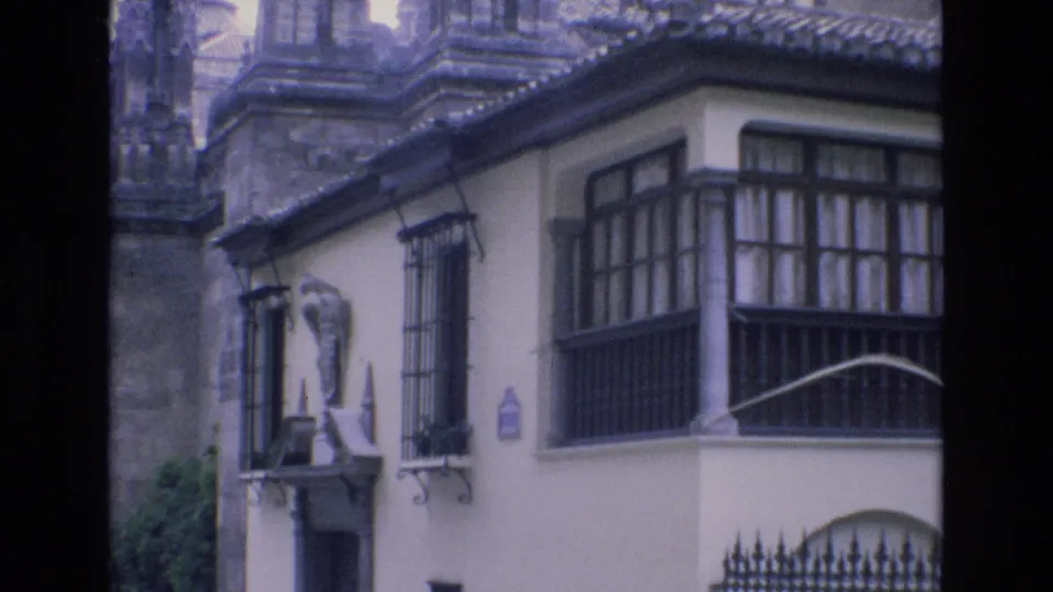 1972 Spain 8mm frame showing a pale urban building with dark balcony windows, wrought-iron grilles, a small exterior statue, and stone architecture behind it.