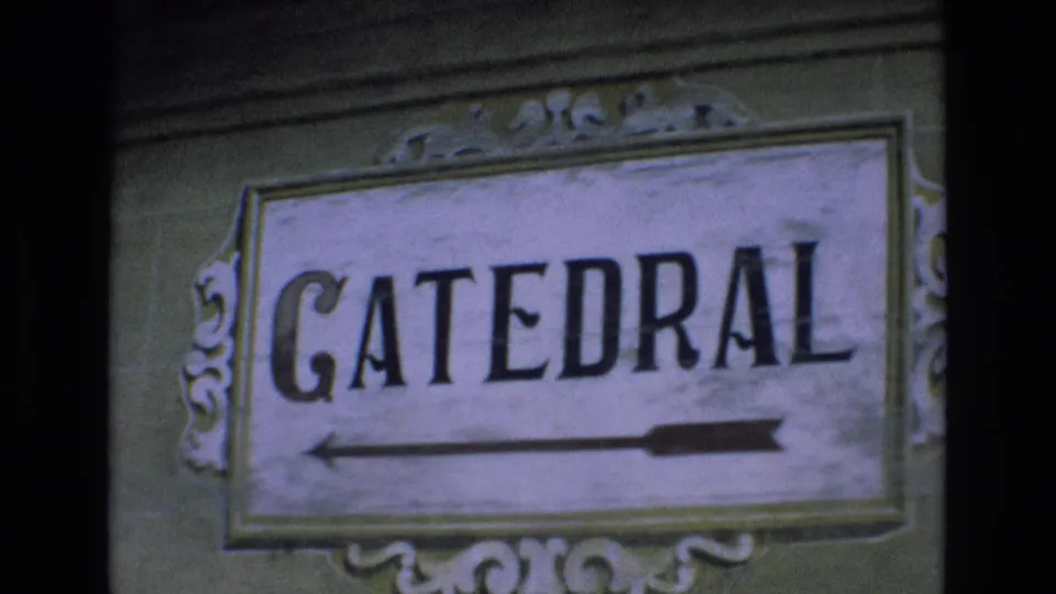 1972 Spain 8mm close-up of a weathered directional sign reading CATEDRAL, with a black arrow pointing left on an ornate rectangular panel.