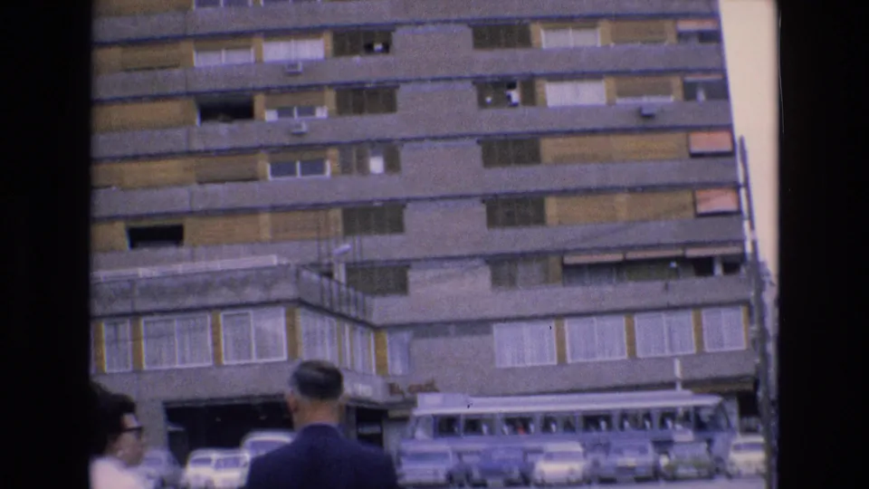 1972 Spain home-movie frame looking toward the lower stories of the building, with parked cars, a coach bus, and two visible people near the foreground.