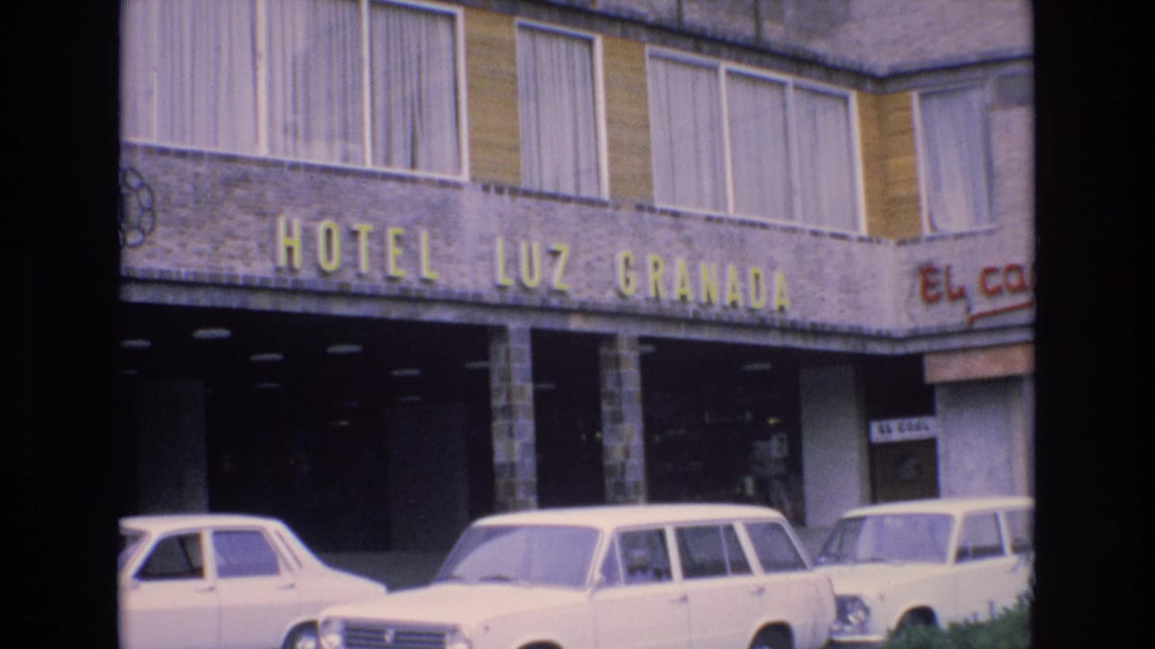 1972 Spain home-movie frame showing the Hotel Luz Granada facade with yellow signage, stone columns, curtained windows, and white cars parked along the street.