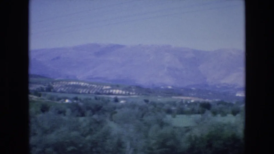 1972 Spain home-movie frame looking over green countryside, rows on a hillside, scattered small structures, and a distant mountain range under pale sky.