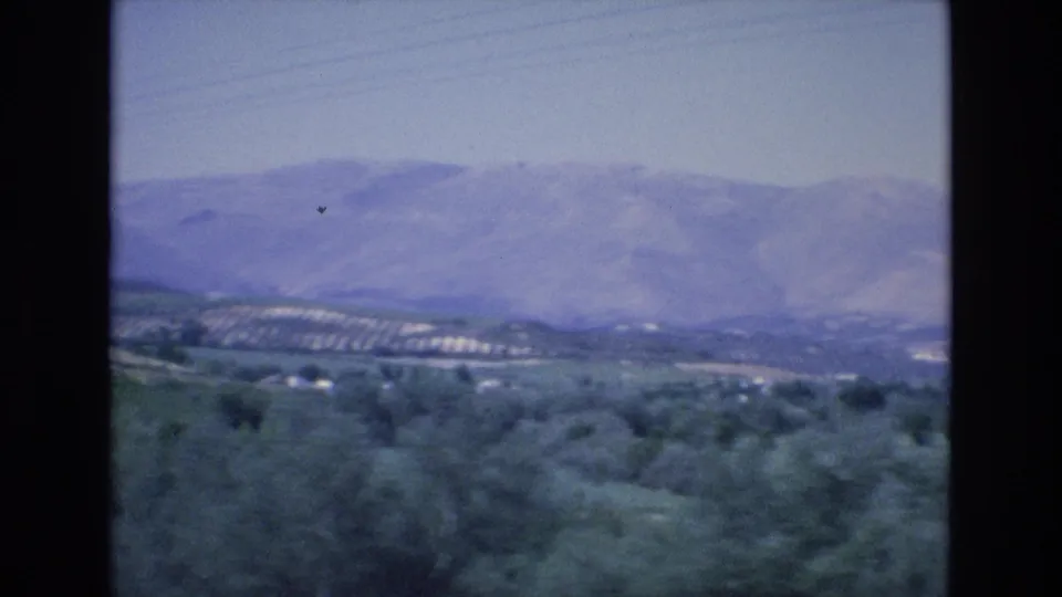 1972 Spain travel-film frame showing trees in the foreground, terraced or patterned hillsides, and a broad hazy mountain range in the distance.