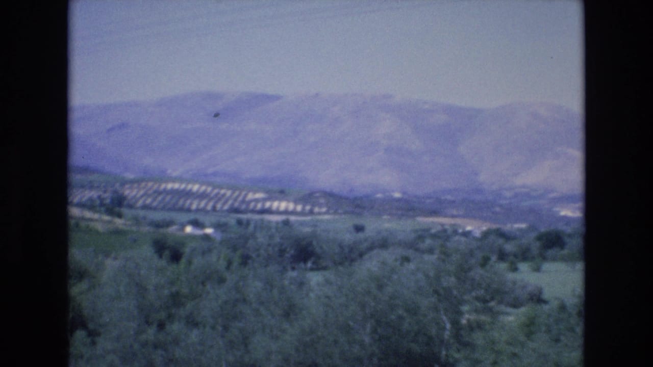 1972 Spain 8mm frame showing hazy mountains, green rural slopes, scattered buildings, and overhead utility wires seen from a moving viewpoint.
