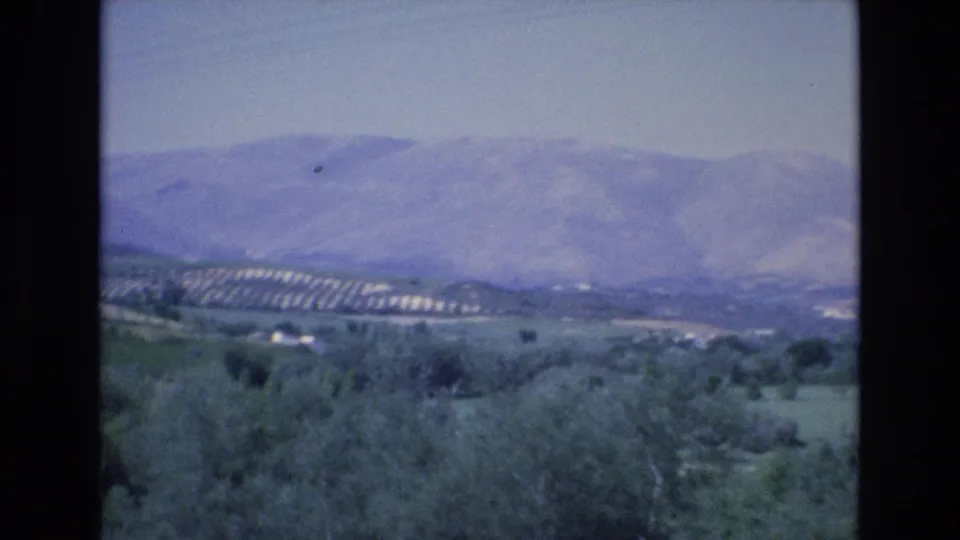 1972 Spain 8mm frame showing hazy mountains, green rural slopes, scattered buildings, and overhead utility wires seen from a moving viewpoint.