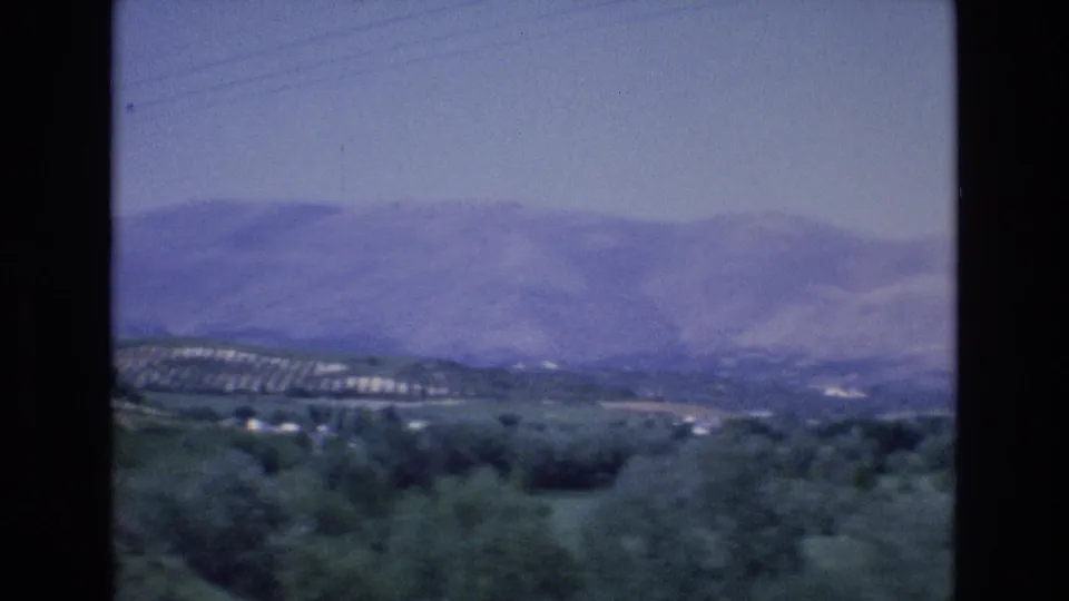 1972 Spain home-movie frame with a broad mountain ridge, green valley vegetation, cultivated slopes, and faint utility lines across the upper sky.
