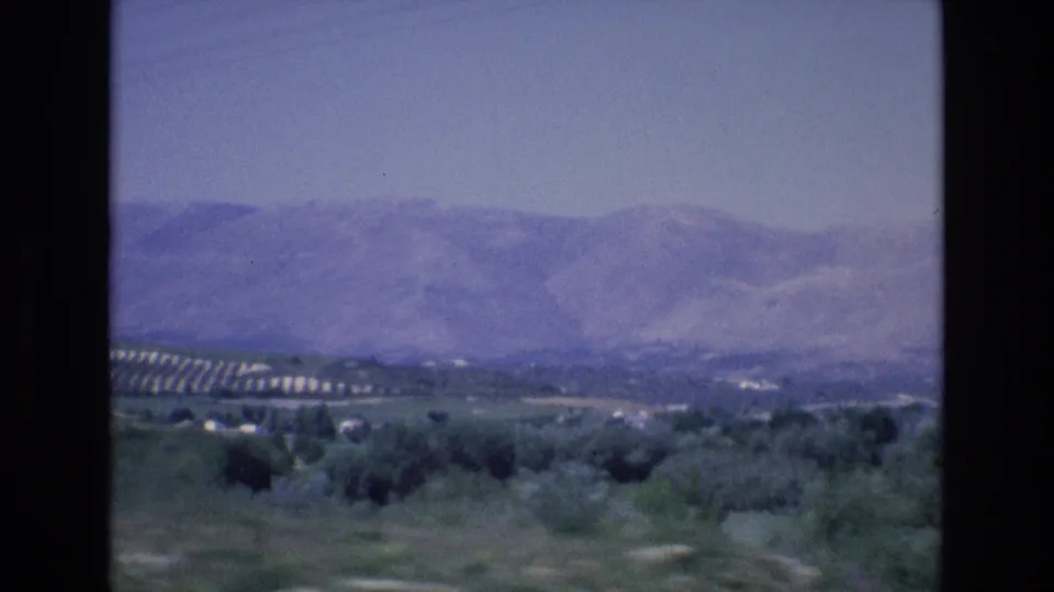 1972 Spain 8mm frame showing pasture, shrubs, distant white buildings, patterned hillside cultivation, and a hazy mountain range.