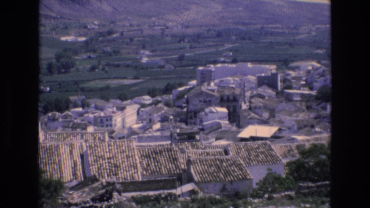 Elevated view over a village with pale buildings, tiled roofs, fields, and mountains beyond.