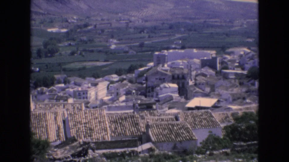 1972 Spain 8mm frame showing an elevated view of a rural village with pale buildings, tiled rooftops, cultivated fields, and mountains in the distance.