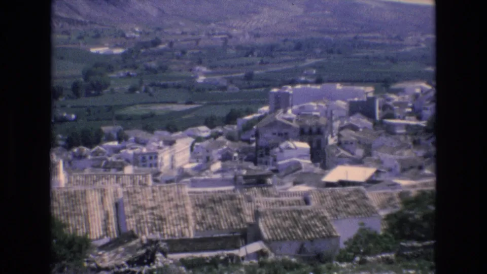 1972 Spain 8mm frame with clustered village buildings and terracotta roof patterns seen from above, set against fields and a mountain valley.