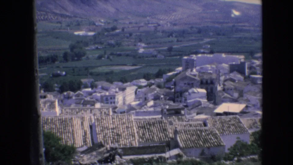 1972 Spain 8mm frame showing the edge of a rural village, tiled roofs, pale structures, green agricultural fields, and hazy mountain terrain beyond.