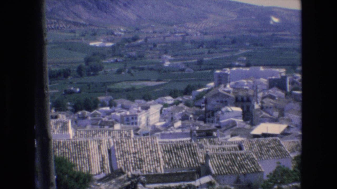 1972 Spain 8mm frame looking across terracotta rooftops and pale village buildings toward green fields and mountain slopes.