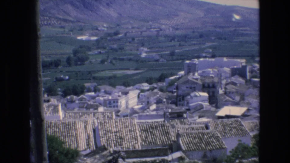 1972 Spain 8mm frame looking across terracotta rooftops and pale village buildings toward green fields and mountain slopes.