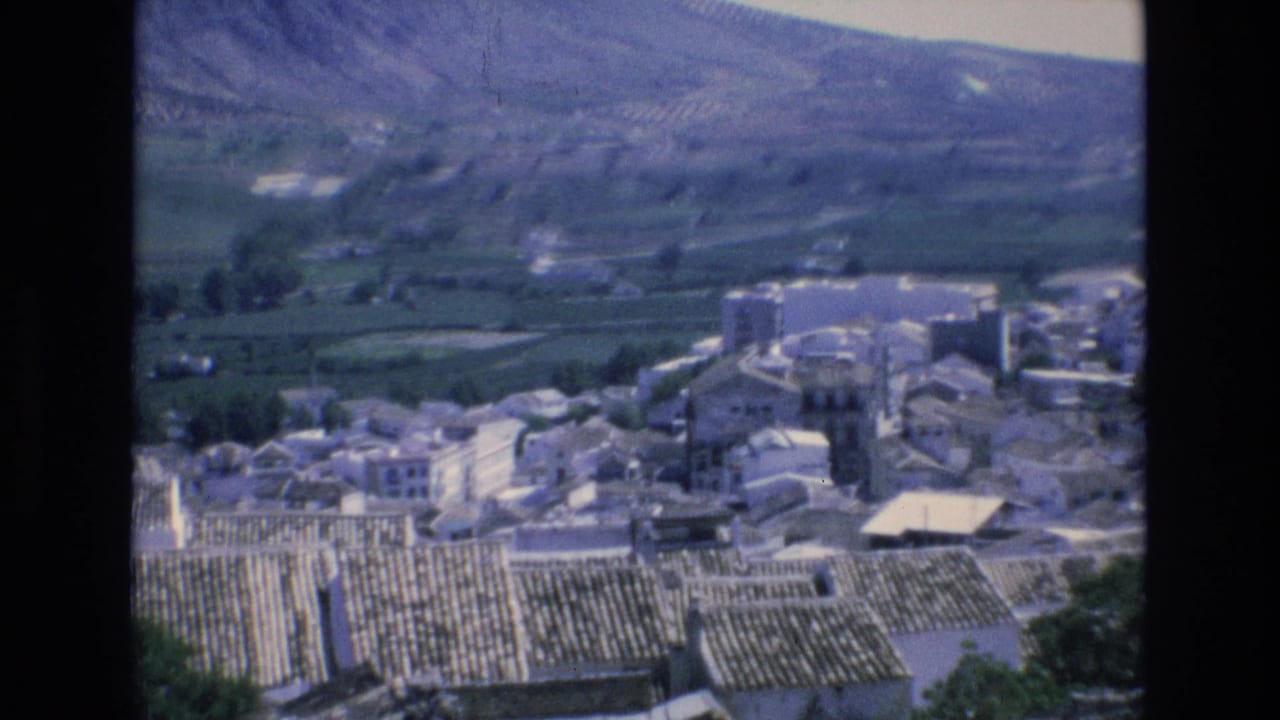 Village rooftops and buildings seen from above with fields and mountains beyond.