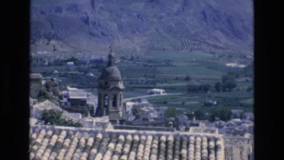 1972 Spain 8mm frame with a church bell tower above tiled rooftops, backed by fields and mountain terrain.