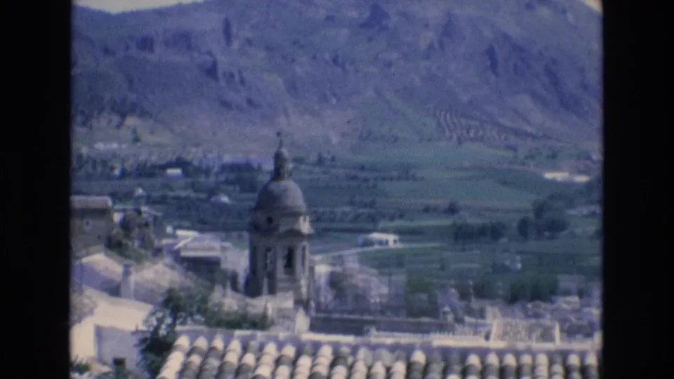 Home-movie frame from 1972 Spain showing a bell tower and roof tiles in the foreground with open fields and mountains beyond.