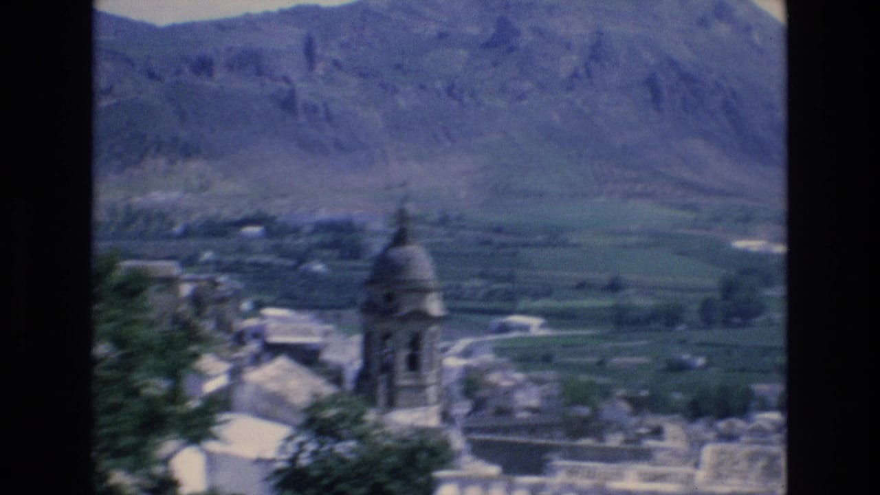 Domed church tower among pale buildings below a mountain valley.