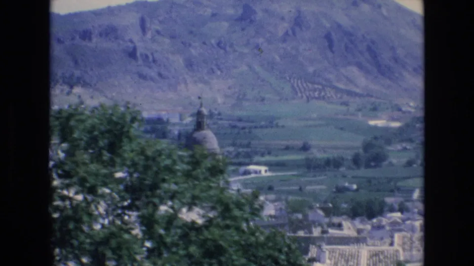 1972 Spain 8mm frame with a leafy tree partly covering a domed church tower, while cultivated valley fields and rocky mountains extend behind it.