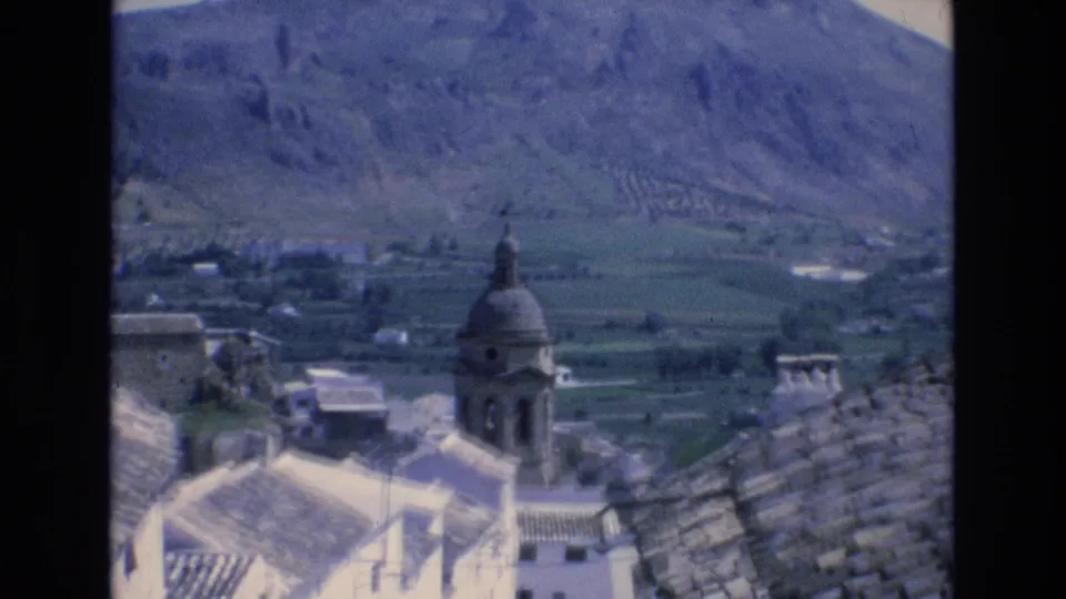 1972 Spain 8mm frame showing a domed church bell tower, whitewashed buildings, tiled rooftops, and agricultural fields at the base of a rugged mountain.