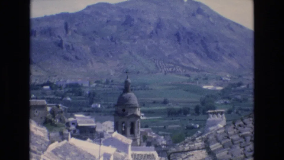 1972 Spain 8mm frame of a domed church tower rising above terracotta rooftops, with a patchwork valley and rocky mountain slope in the distance.