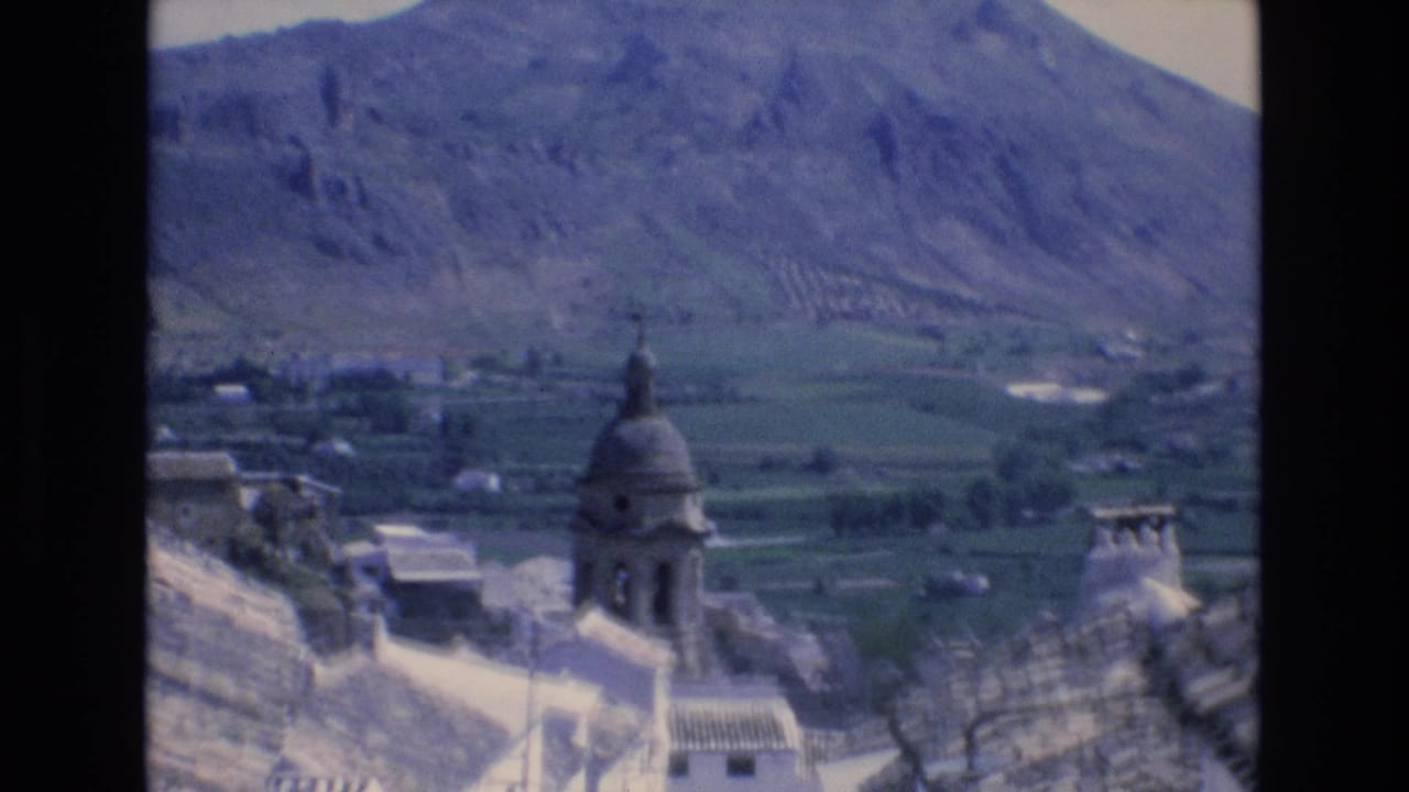 1972 Spain 8mm frame showing a church tower rising above white village rooftops, with cultivated valley fields and rugged mountains in the distance.