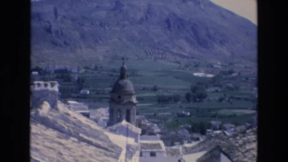 1972 Spain home-movie view of a church dome and tiled village rooftops from an elevated position, backed by fields and rocky mountain terrain.