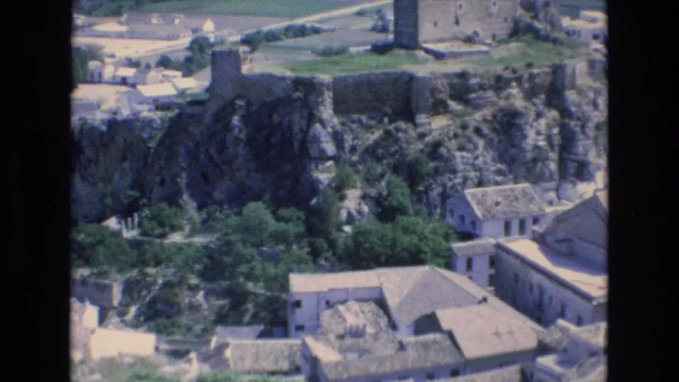 1972 Spain 8mm frame showing stone ruins on a rocky outcrop above white village buildings, trees, and cultivated land.