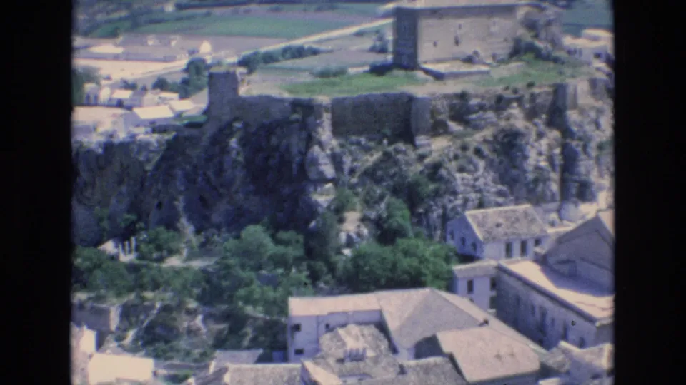 1972 Spain home-movie frame with rocky hillside ruins, trees, pale village buildings, and valley fields seen from an elevated viewpoint.