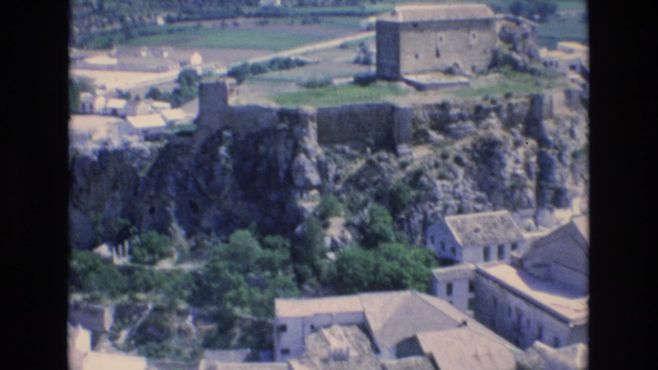 1972 Spain 8mm frame showing stone fortress-like ruins on a rocky outcrop above clustered buildings and distant agricultural fields.